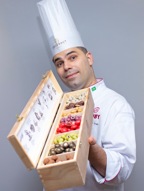 Chef Abner Ivan segurando uma caixa de madeira com sete variedades de drágeas separadas por compartimento.
